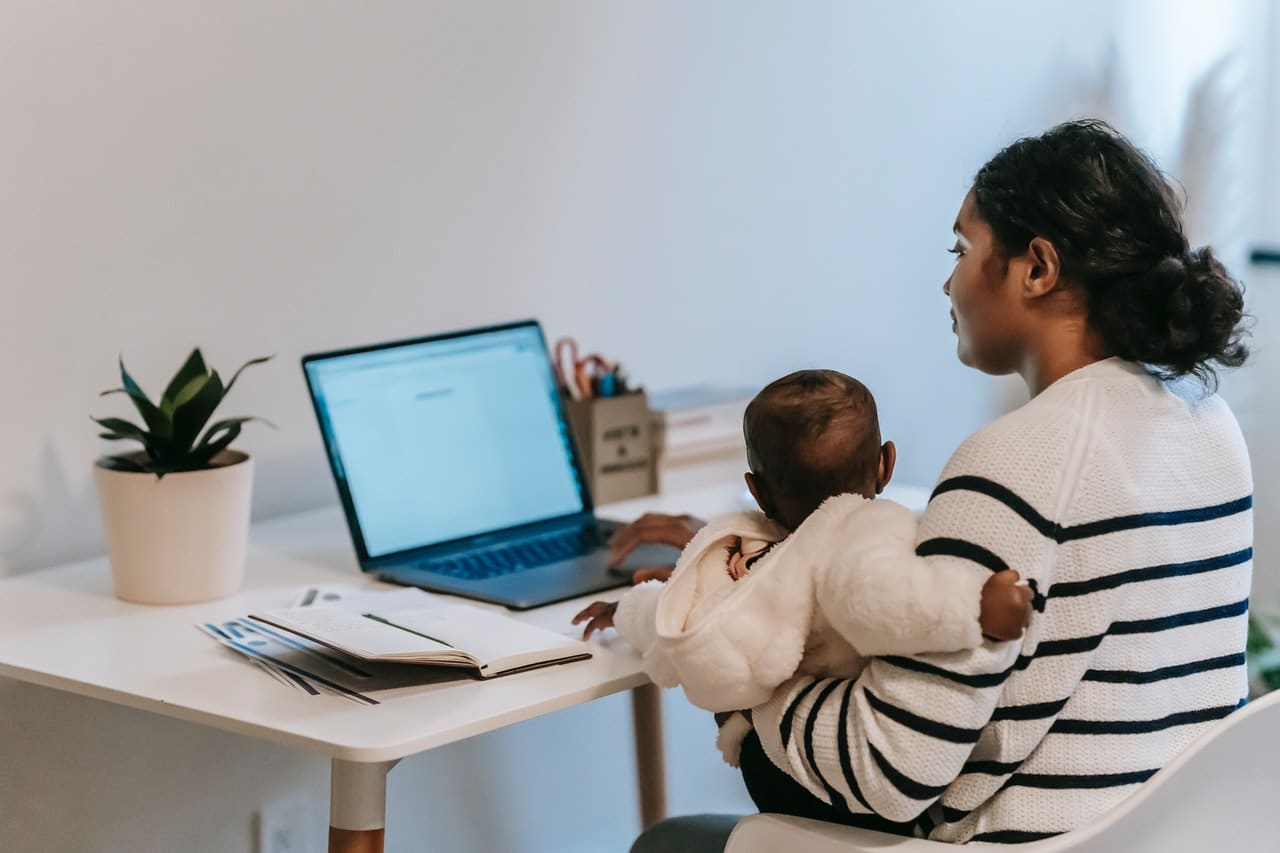 A mother holding her baby while sitting at a computer representing how the pandemic has made it difficult for new parents to get Employment Insurance during parental leave