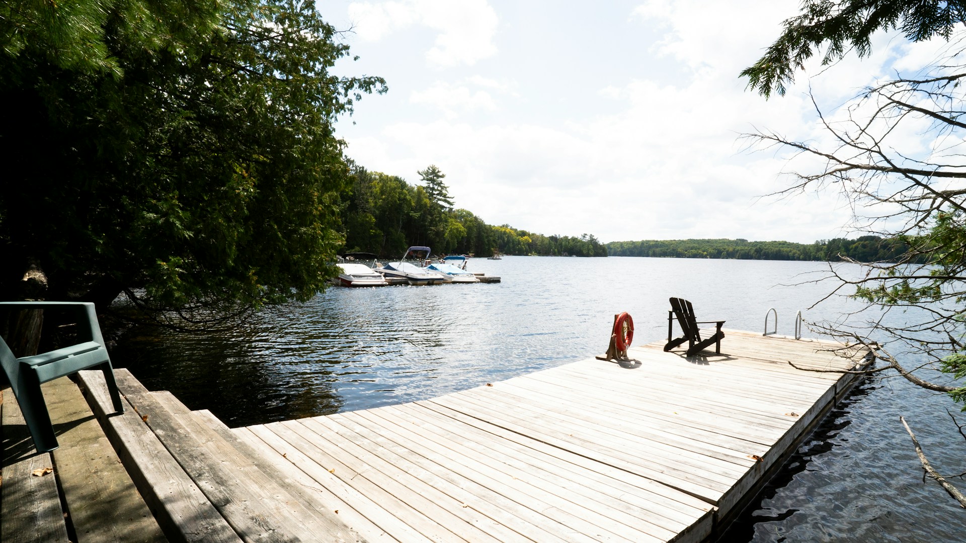 Wooden dock extending into a calm lake with boats moored in the distance, two chairs and a life preserver on the dock, and trees lining the shore under a cloudy sky.