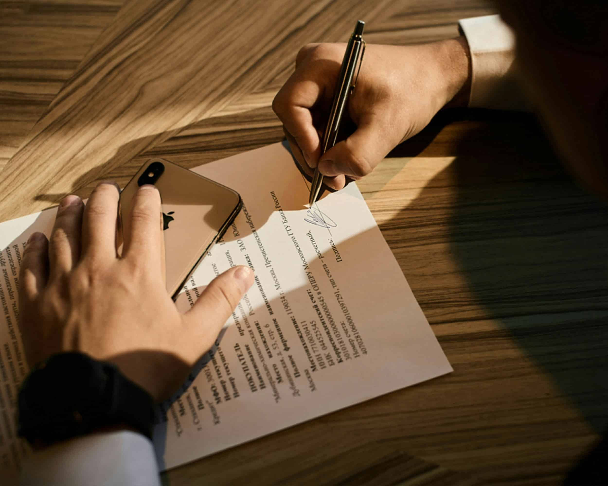 Close-up of a person's hands signing a document on a wooden desk with a gold smartphone nearby, illustrating the signing of a legal agreement, potentially related to pre-incorporation contracts in Ontario.