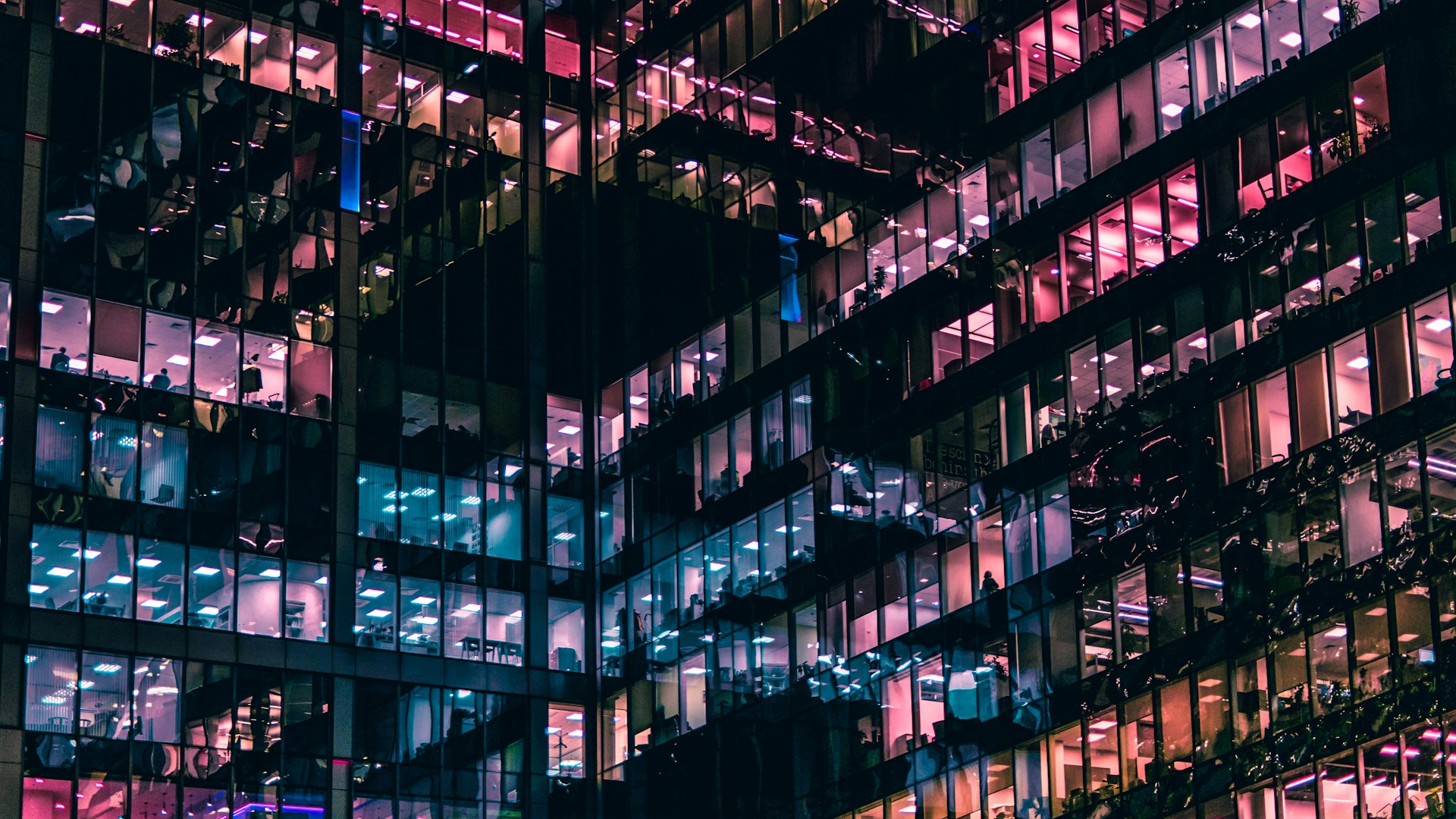 The side of a glass office building at night with the windows lit up by pastel pink, purple, blue, and orange light, showing the people inside, representing the Competition Act amendments.