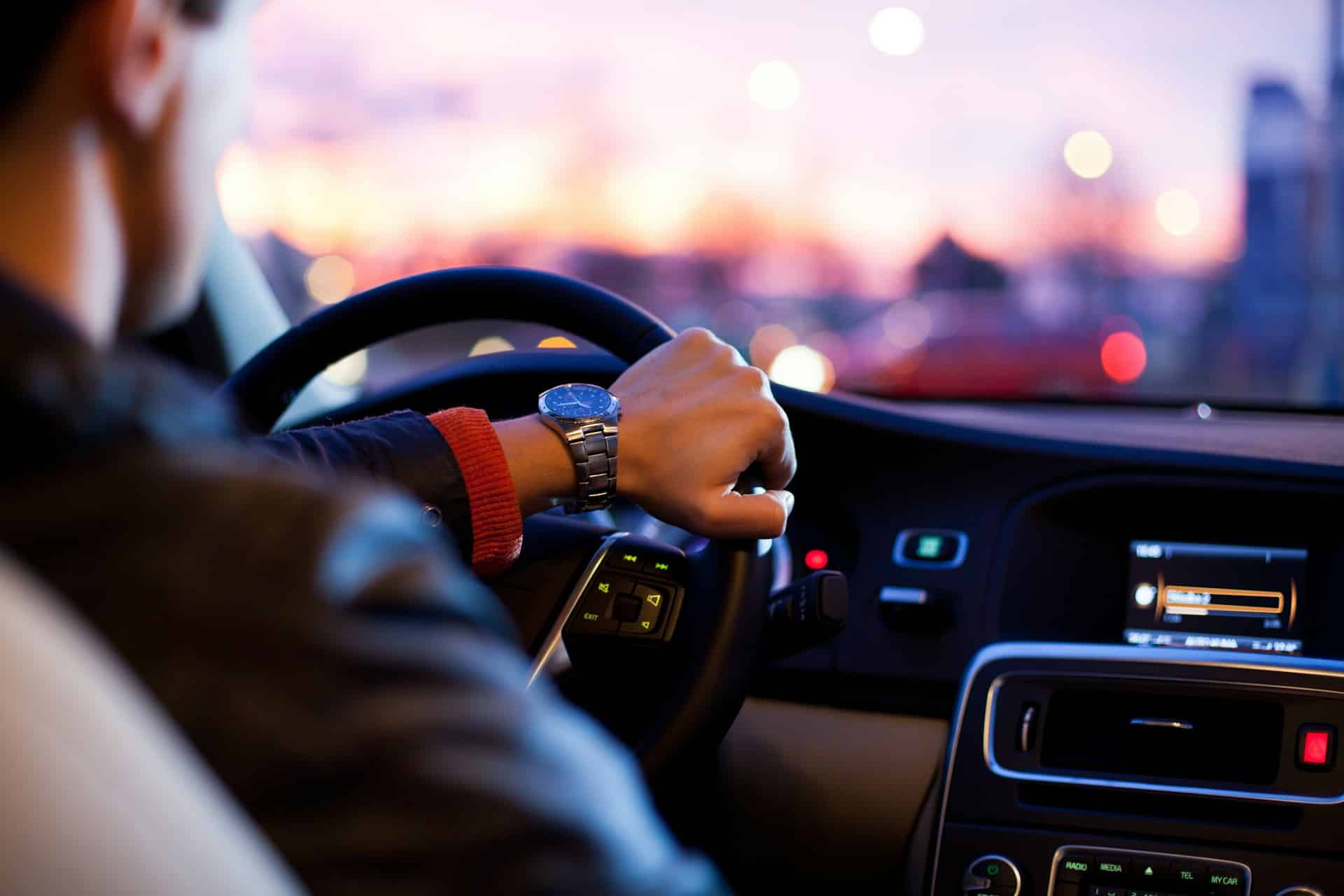 A man driving his car is shown from behind and in profile with the city at dusk blurred through the windshield, symbolizing a motor vehicle accident like the one featured and the denial of SABS.