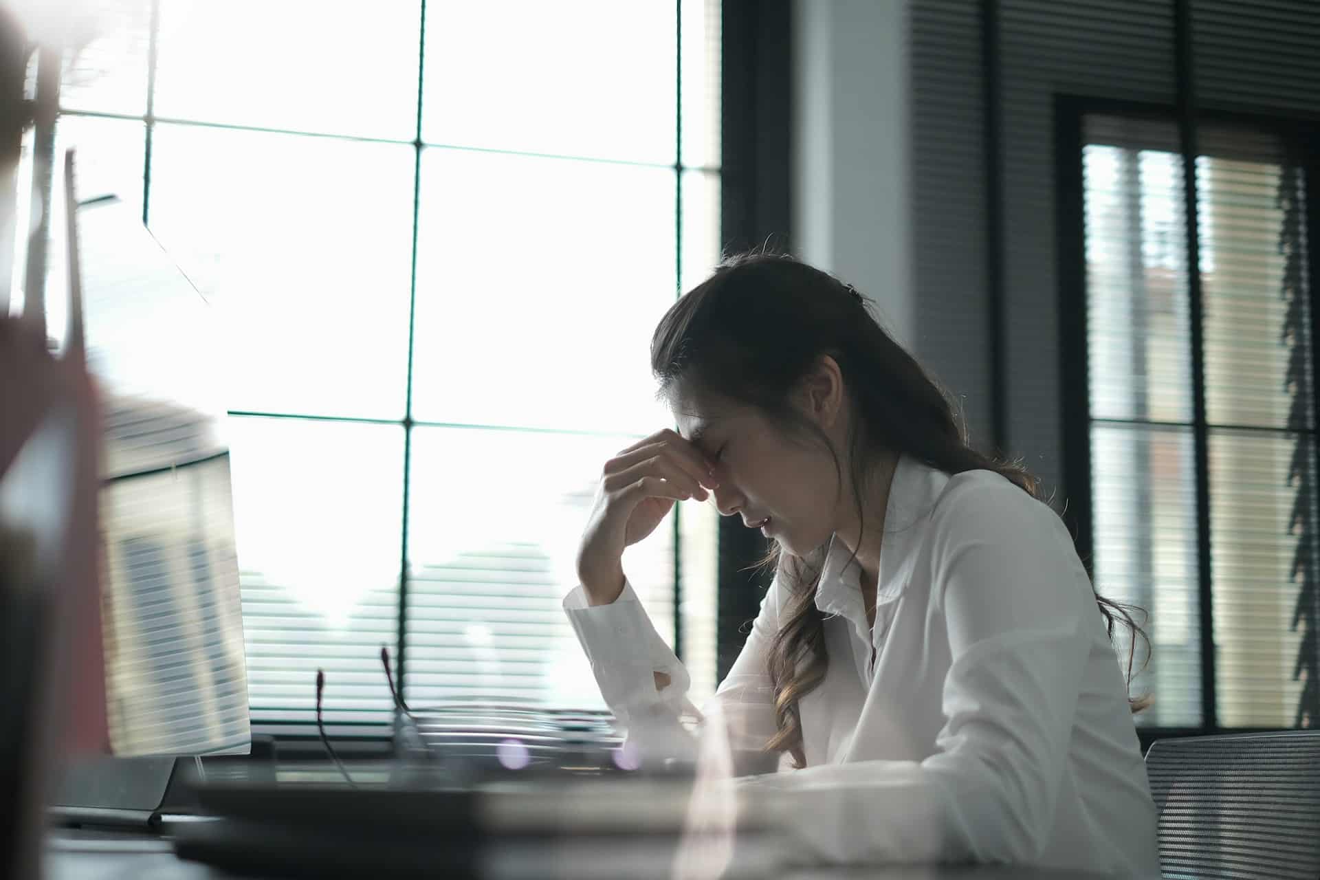 A woman sitting at a desk with her eyes closed and pinching the bridge of her nose after removing her glasses and setting them on the desk next to a blurred computer, representing toxic workplaces in Ontario.