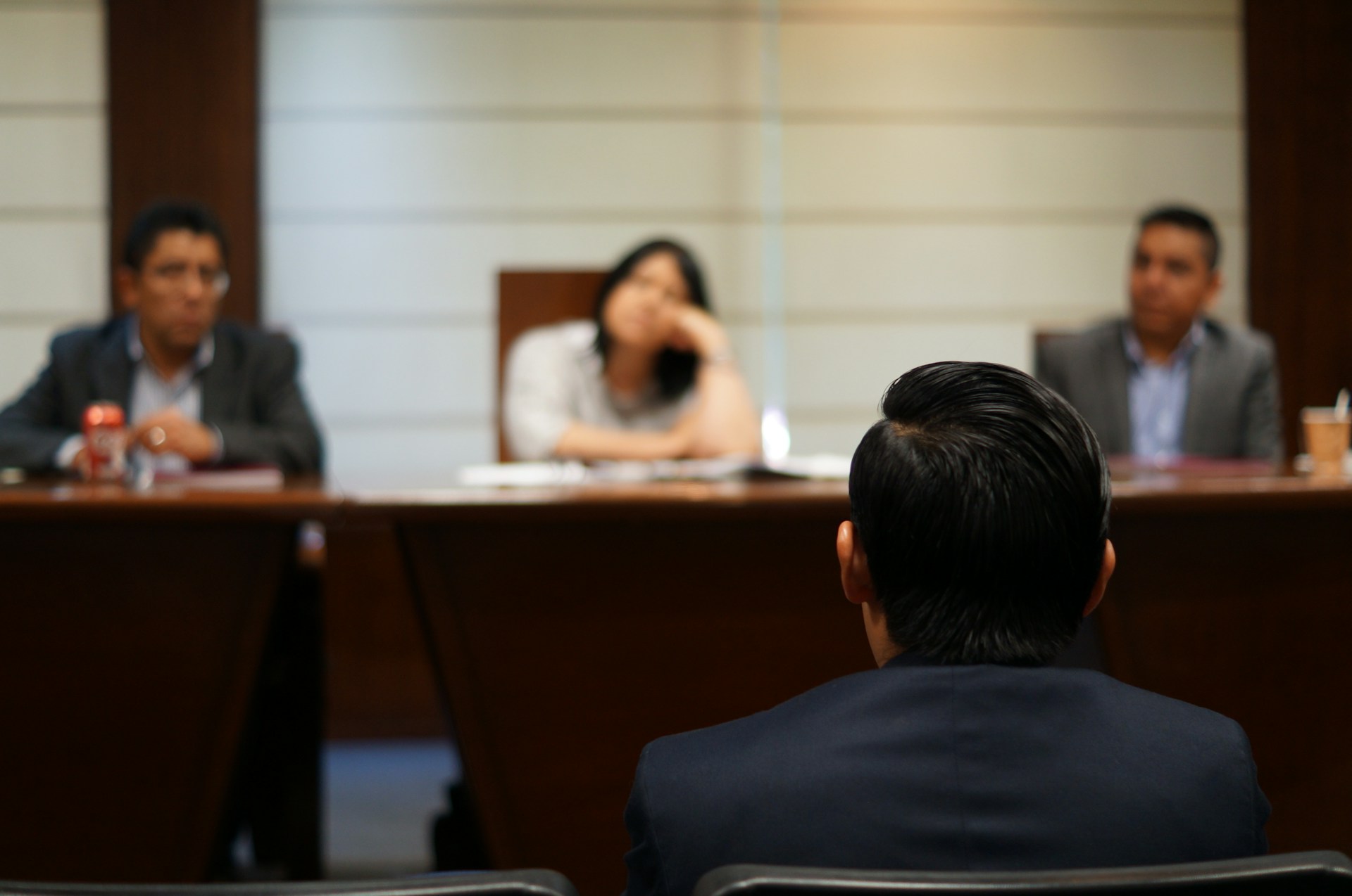 A man sits before a blurred pannel, two men and a woman, seated behind a long wooden desk, representing social benefits tribunal.