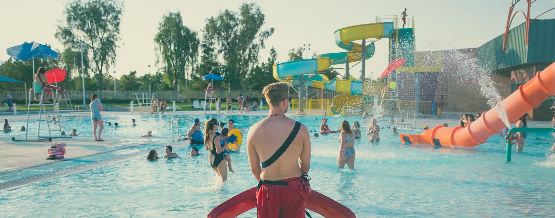 A lifeguard stands at the edge of a water park pool with multiple slides, watching over the families playing in the water, representing Ontario beach and water park injuries.